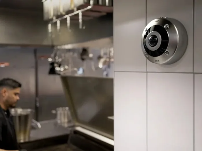 Cámara de vigilancia redonda instalada en una pared de azulejos blancos dentro de una cocina profesional con un trabajador al fondo.