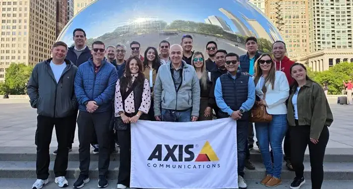 Grupo de personas sonrientes sosteniendo una bandera de Axis Communications frente a una gran estructura esférica reflectante.