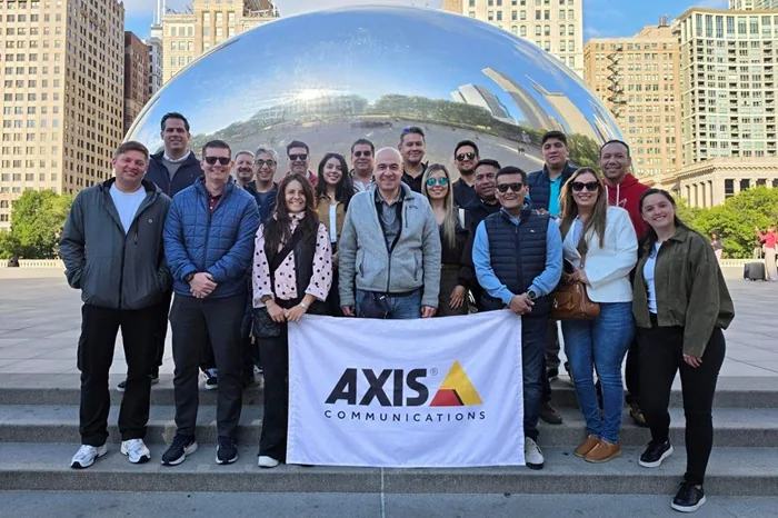 Grupo de personas sonrientes sosteniendo una bandera de Axis Communications frente a una gran estructura esférica reflectante.