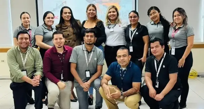 Grupo diverso de profesionales sonriendo y posando juntos en una sala de reuniones corporativa con una pantalla al fondo.