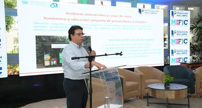 Hombre hablando ante una pantalla con información sobre simbiosis industrial y casos de éxito en una conferencia.