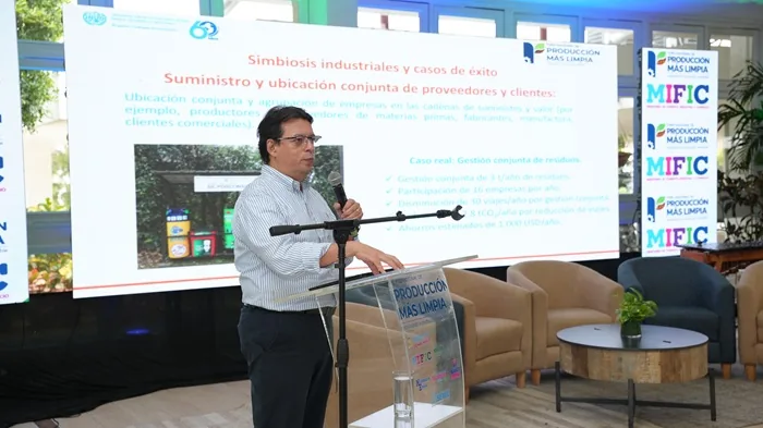 Hombre hablando ante una pantalla con información sobre simbiosis industrial y casos de éxito en una conferencia.