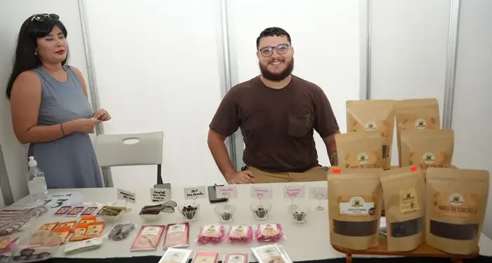 Dos emprendedores en un stand de feria, mostrando productos de cacao y chocolate. La mesa exhibe barras, nibs y muestras en empaques artesanales.