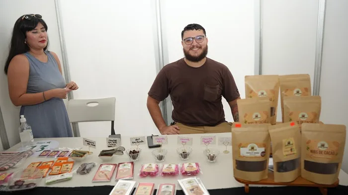 Dos emprendedores en un stand de feria, mostrando productos de cacao y chocolate. La mesa exhibe barras, nibs y muestras en empaques artesanales.