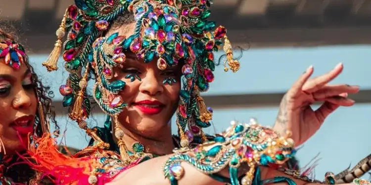Mujer sonriente con un elaborado tocado y traje decorado con piedras preciosas de colores verde, azul y rosa en un desfile.