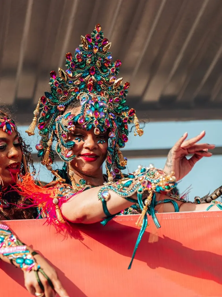 Mujer sonriente con un elaborado tocado y traje decorado con piedras preciosas de colores verde, azul y rosa en un desfile.