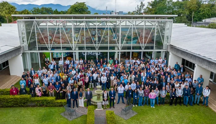 Vista aérea frontal de un numeroso grupo de profesionales posando frente a la entrada de un edificio corporativo moderno con mural