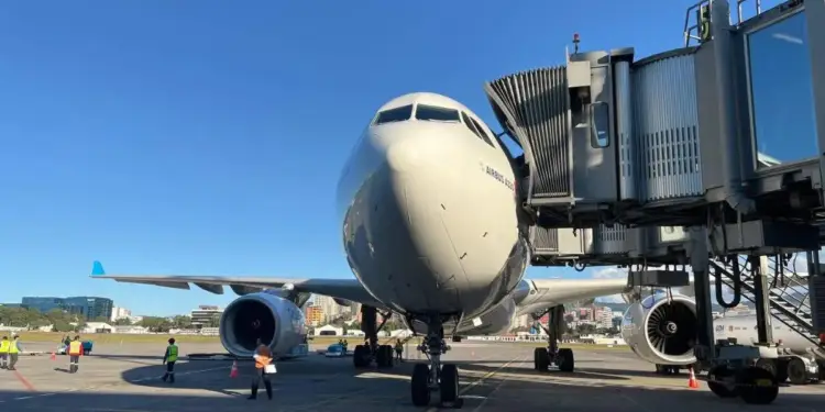 Vista frontal de un avión comercial Airbus A330 estacionado en la pista junto a una manga de abordaje y personal técnico.