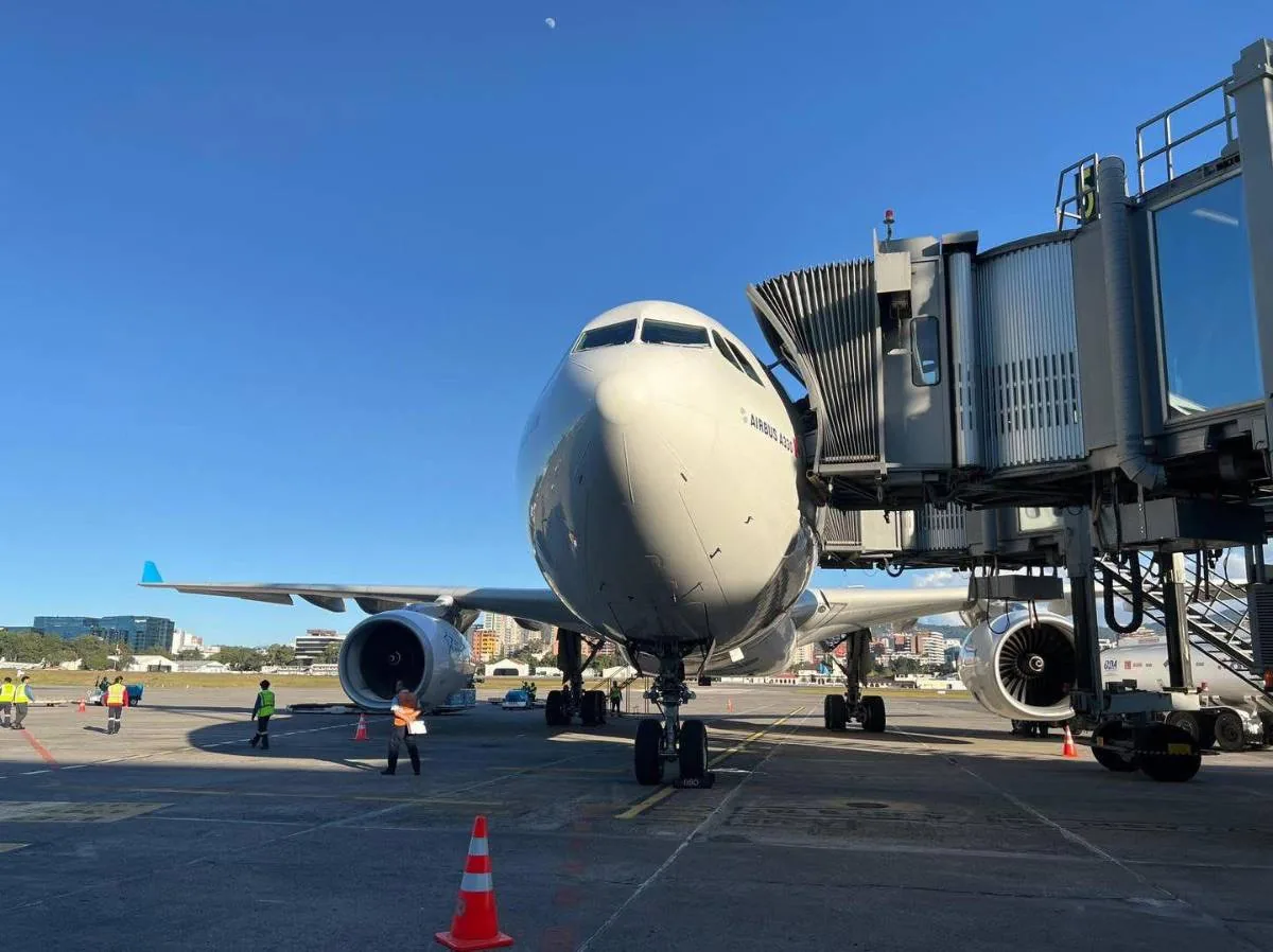 Vista frontal de un avión comercial Airbus A330 estacionado en la pista junto a una manga de abordaje y personal técnico.