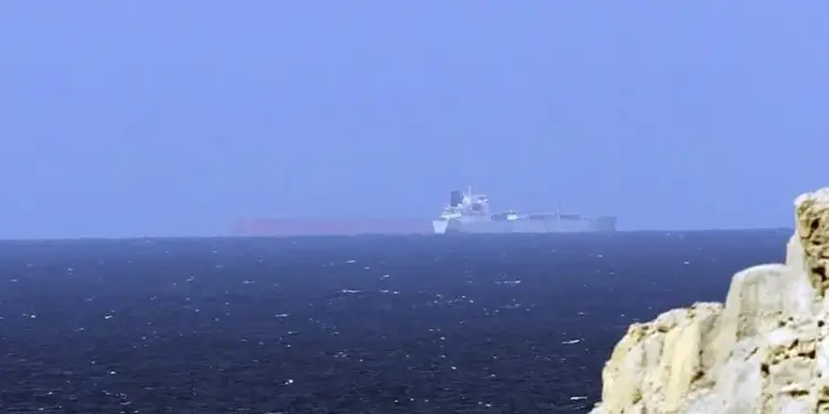Un gran barco de carga navegando en el mar abierto con formaciones rocosas en primer plano a la derecha.