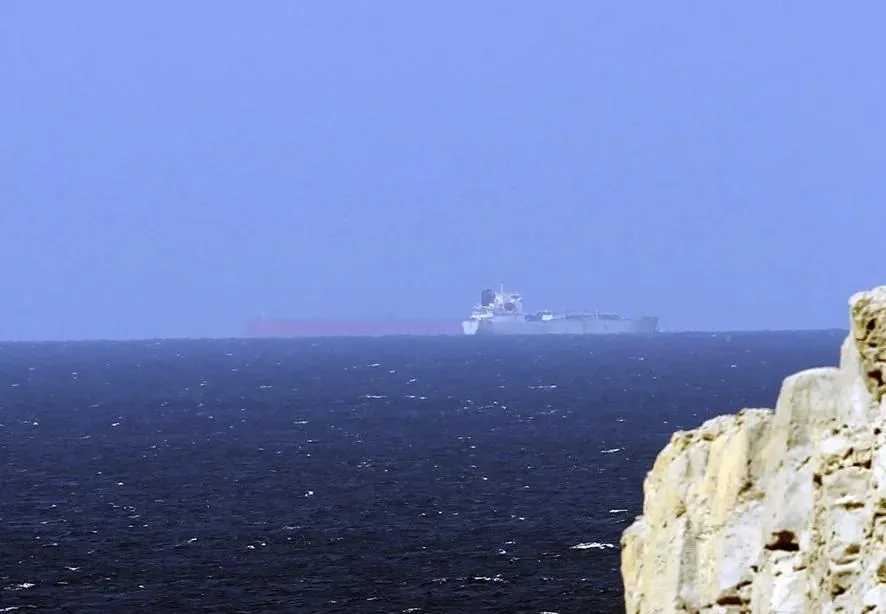 Un gran barco de carga navegando en el mar abierto con formaciones rocosas en primer plano a la derecha.