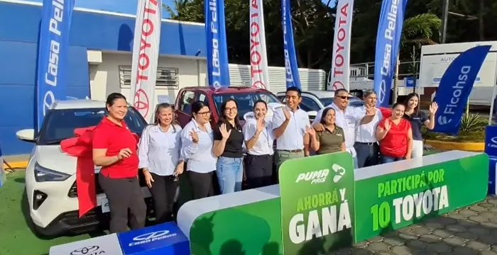 Grupo de personas sonrientes posando frente a vehículos nuevos junto a banderas corporativas y un letrero promocional.