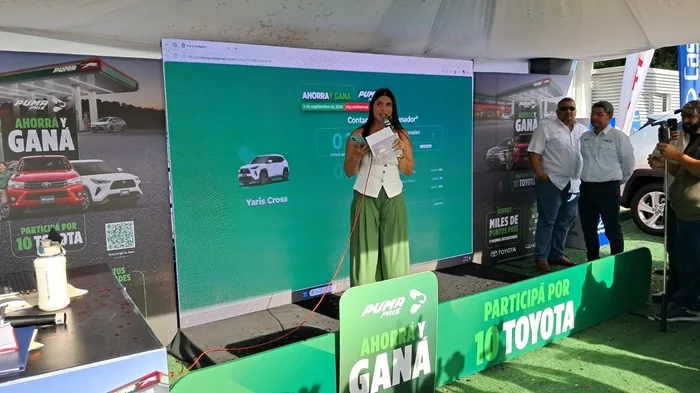 Mujer hablando con micrófono en un evento corporativo frente a una pantalla grande con publicidad.
