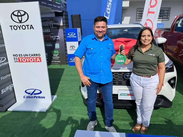 Un hombre y una mujer sonriendo junto a un automóvil blanco nuevo, sosteniendo una llave en un evento promocional.