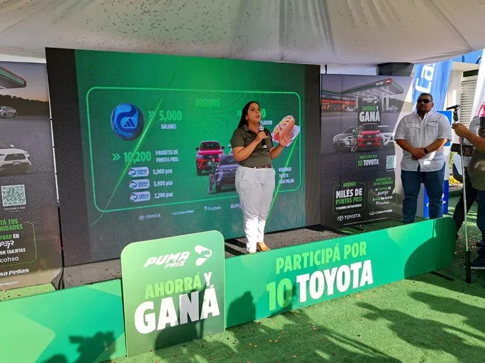 Una mujer habla por un micrófono frente a una pantalla gigante con gráficos de una campaña publicitaria en un evento al aire libre.