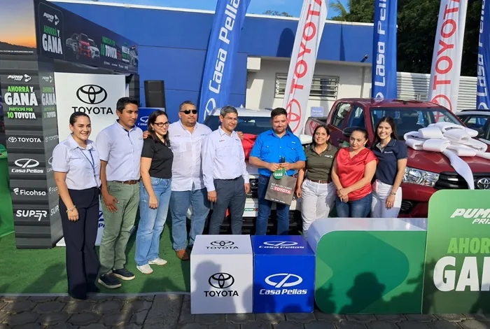 Grupo de nueve personas posando sonrientes frente a vehículos y carteles promocionales con logotipos corporativos.