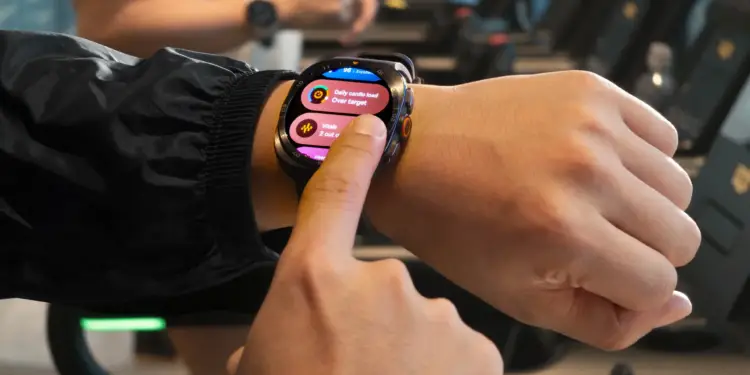 Primer plano de una persona usando un smartwatch en su muñeca, tocando la pantalla que muestra métricas de salud y carga cardiovascular. Al fondo, equipo de gimnasio borroso.