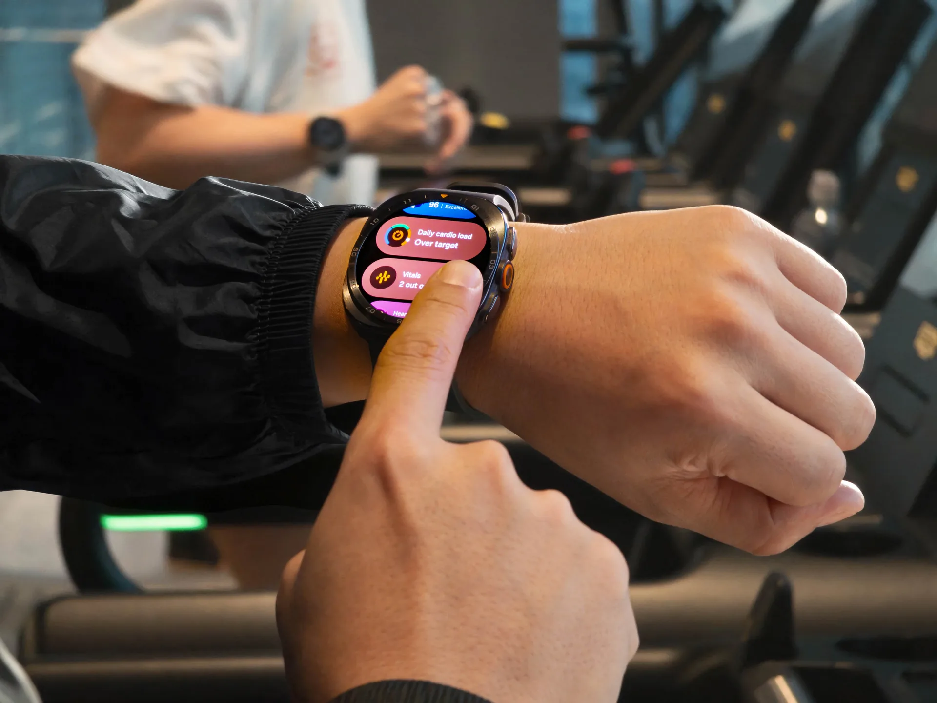Primer plano de una persona usando un smartwatch en su muñeca, tocando la pantalla que muestra métricas de salud y carga cardiovascular. Al fondo, equipo de gimnasio borroso.