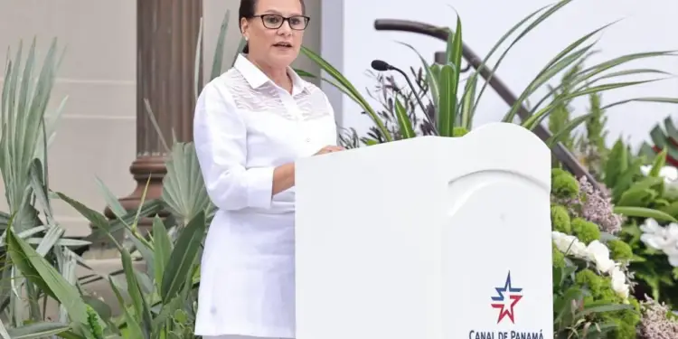 Mujer con gafas y vestimenta blanca hablando en un podio blanco con el logo 'CANAL DE PANAMÁ'. Fondo con vegetación y columna.