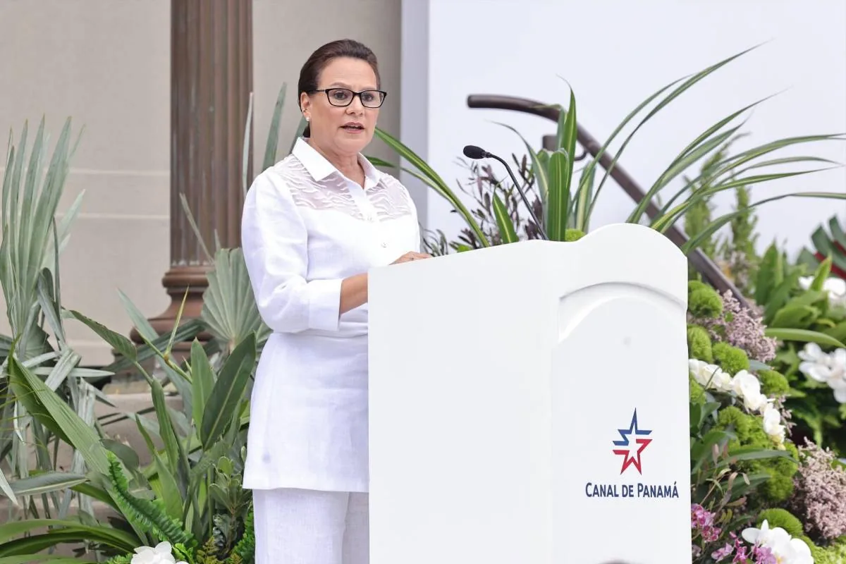 Mujer con gafas y vestimenta blanca hablando en un podio blanco con el logo 'CANAL DE PANAMÁ'. Fondo con vegetación y columna.