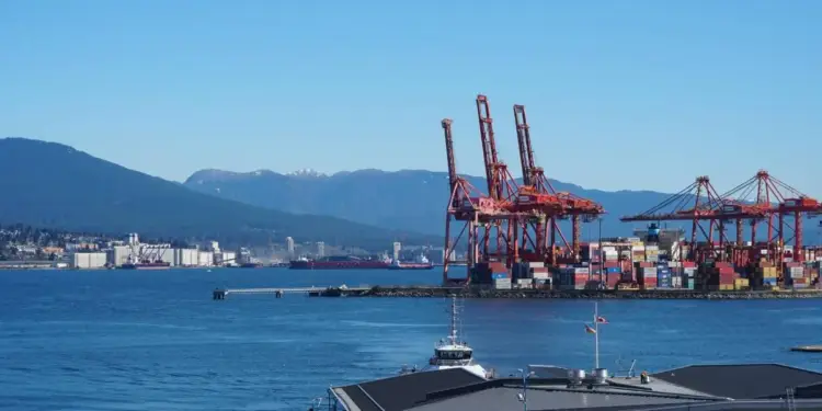 Vista panorámica de un puerto comercial con grúas rojas y contenedores frente al mar bajo un cielo despejado.