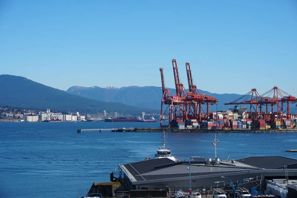 Vista panorámica de un puerto comercial con grúas rojas y contenedores frente al mar bajo un cielo despejado.