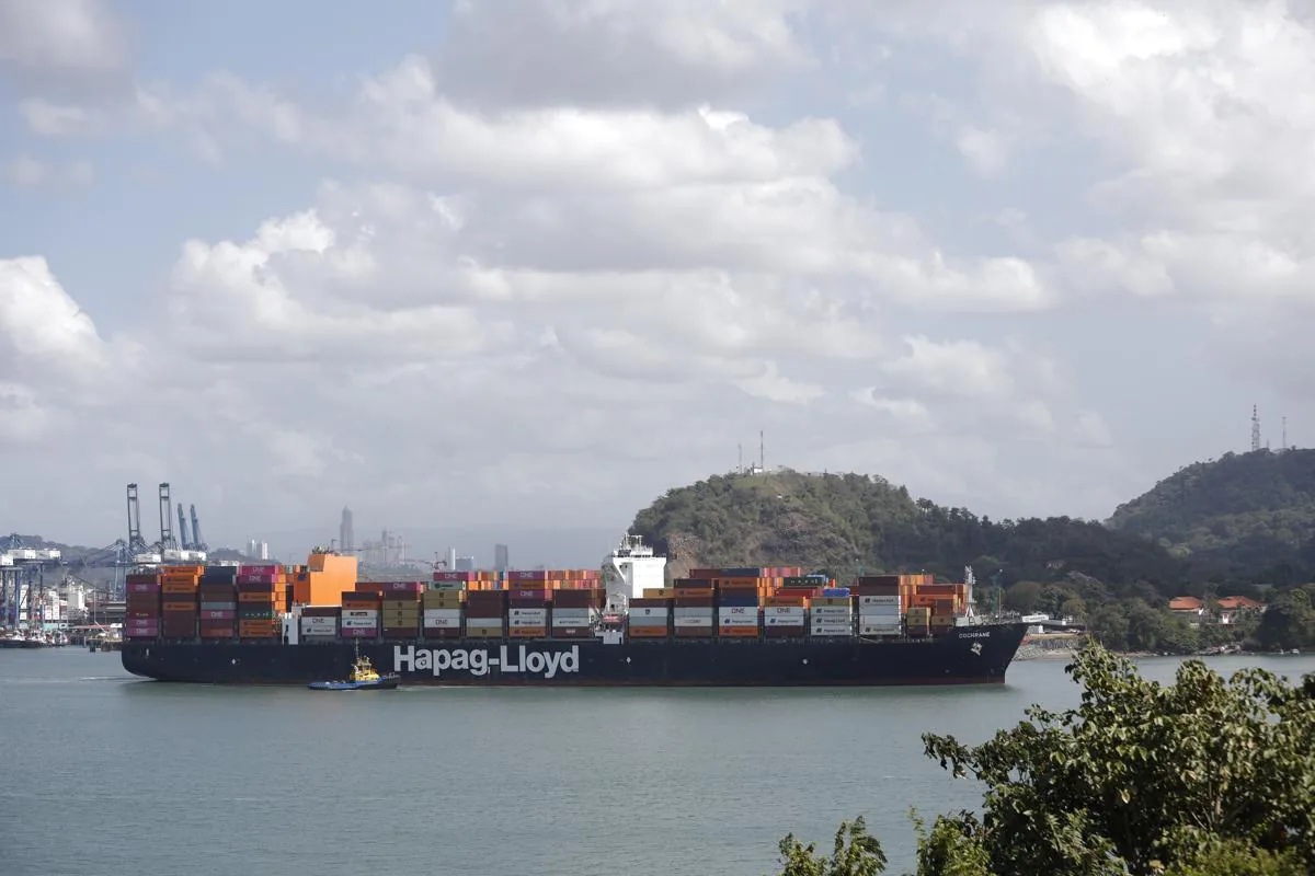 Un gran buque portacontenedores de Hapag-Lloyd navegando en el mar con contenedores de colores y un pequeño remolcador.