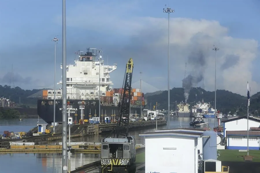 Gran buque portacontenedores con texto 'CHINA SINGAPORE' en su casco, navegando por un canal. Se ven una locomotora, grúa, humo y colinas.