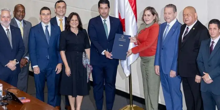 Once profesionales, hombres y mujeres, en trajes formales intercambian un documento azul con la palabra 'PLENO' en una sala oficial con una bandera.