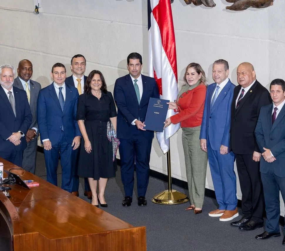 Once profesionales, hombres y mujeres, en trajes formales intercambian un documento azul con la palabra 'PLENO' en una sala oficial con una bandera.