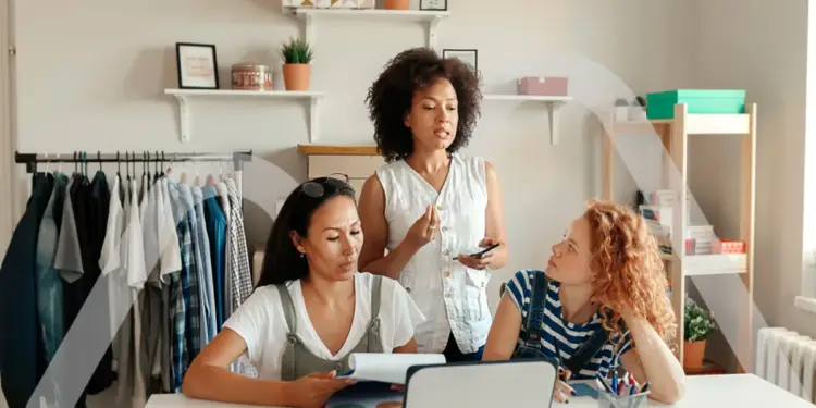 Tres mujeres diversas trabajando juntas en una oficina o taller de moda, revisando documentos y usando una laptop, con ropa colgada en un perchero al fondo.