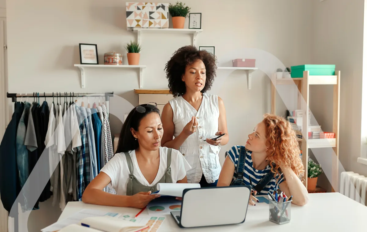 Tres mujeres diversas trabajando juntas en una oficina o taller de moda, revisando documentos y usando una laptop, con ropa colgada en un perchero al fondo.