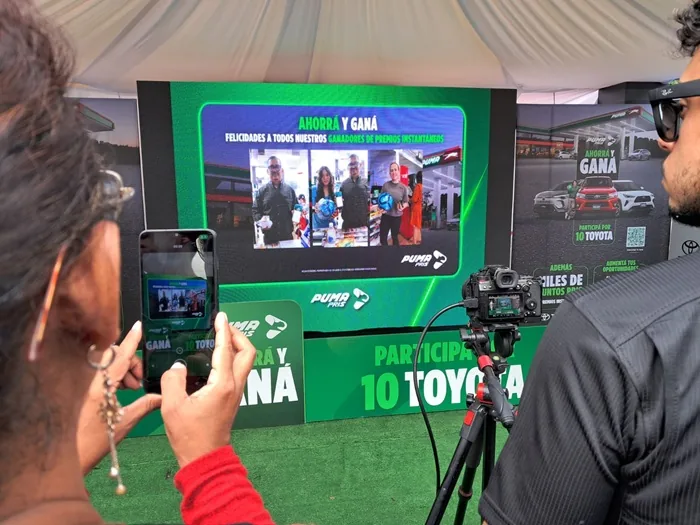 Persona grabando con un teléfono móvil una pantalla que muestra una presentación promocional con imágenes de ganadores.