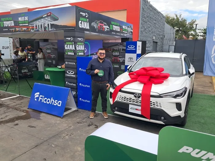 Hombre sonriente junto a un automóvil blanco decorado con un gran lazo rojo en evento promocional corporativo.