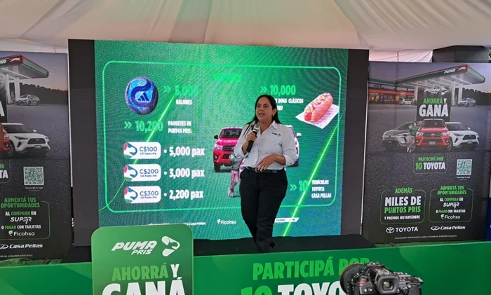 Una mujer con camisa blanca y pantalón negro sostiene un micrófono frente a una pantalla LED verde con gráficos y números.