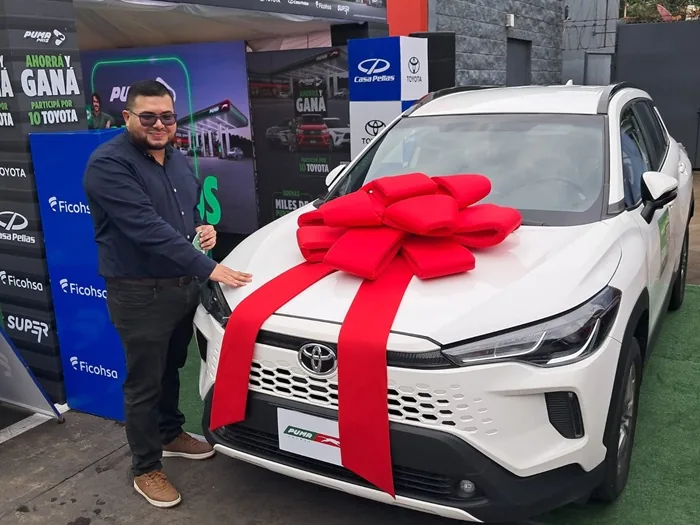 Hombre sonriente junto a un automóvil blanco decorado con un gran lazo rojo en un evento empresarial.