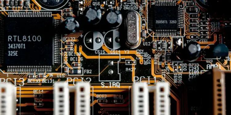 Primer plano de una placa de circuito impreso (PCB) con microchips, condensadores, resistencias y trazas doradas. Se aprecian ranuras de expansión PCI.