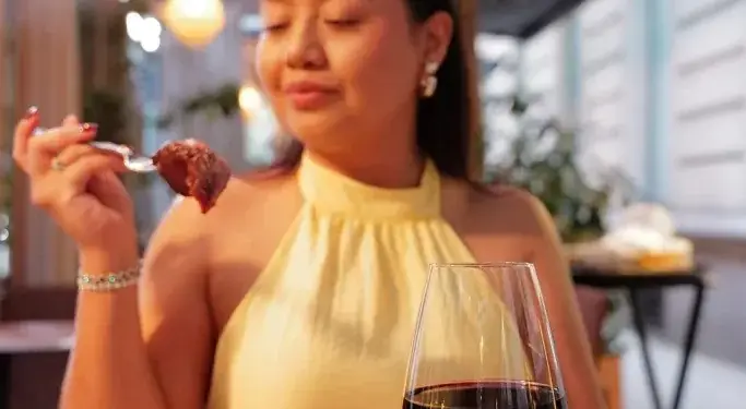 Una mujer con blusa amarilla sostiene un tenedor con un trozo de carne frente a un plato de carne asada y vino tinto.