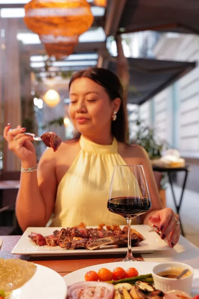 Una mujer con blusa amarilla sostiene un tenedor con un trozo de carne frente a un plato de carne asada y vino tinto.
