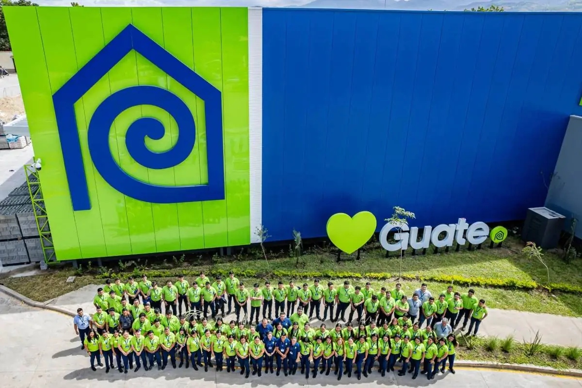 Gran grupo de empleados posando frente a la fachada azul y verde de un edificio comercial con un logotipo de una casa.