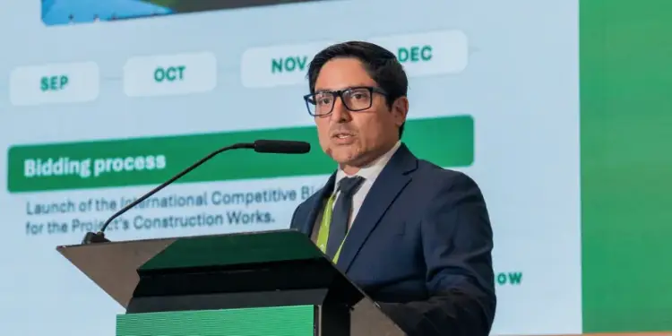 Hombre de traje oscuro y gafas hablando en un atril frente a una pantalla con gráficos en una conferencia empresarial.