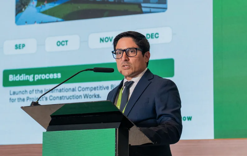 Hombre de traje oscuro y gafas hablando en un atril frente a una pantalla con gráficos en una conferencia empresarial.