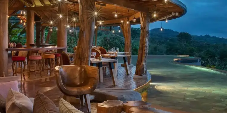 Terraza de restaurante con estructura de madera, iluminación colgante y vista a la naturaleza al atardecer.