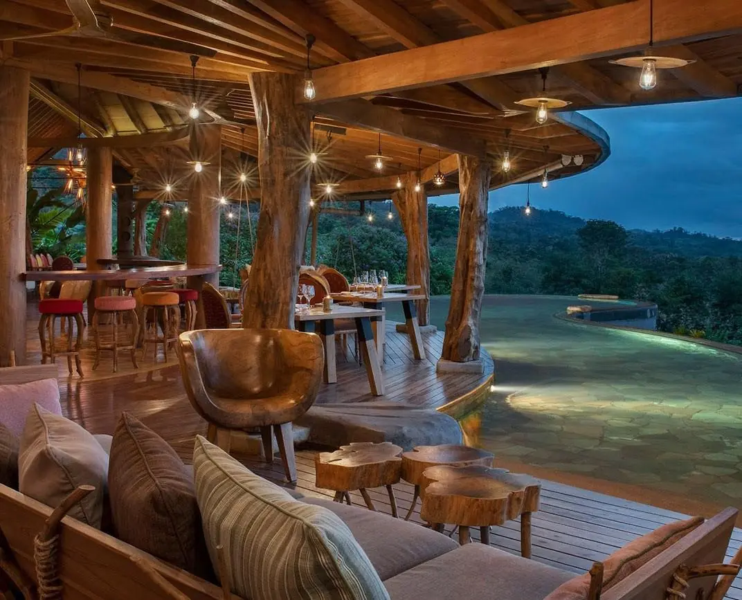 Terraza de restaurante con estructura de madera, iluminación colgante y vista a la naturaleza al atardecer.