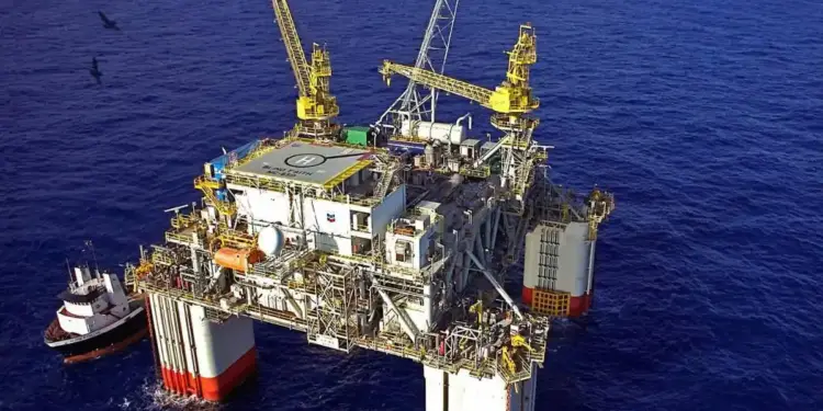 Plataforma petrolera flotante en alta mar con grúas amarillas, helipuerto y un barco de apoyo al lado.