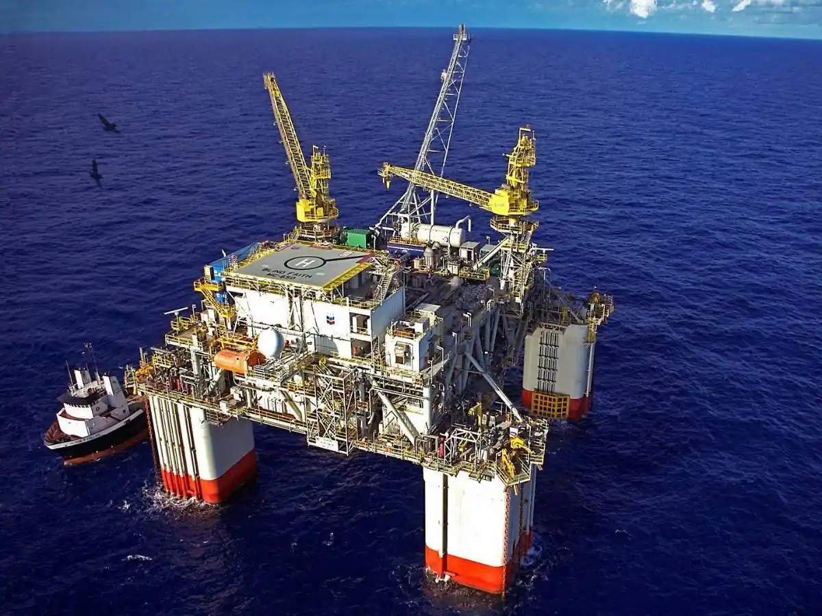 Plataforma petrolera flotante en alta mar con grúas amarillas, helipuerto y un barco de apoyo al lado.