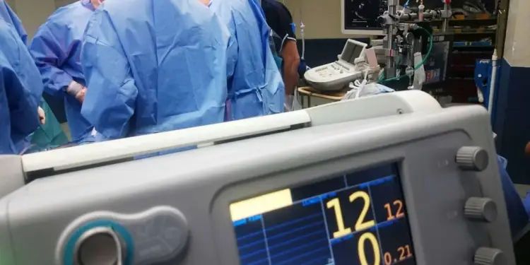 Primer plano de un monitor médico con pantallas digitales en funcionamiento dentro de un quirófano con cirujanos de fondo.