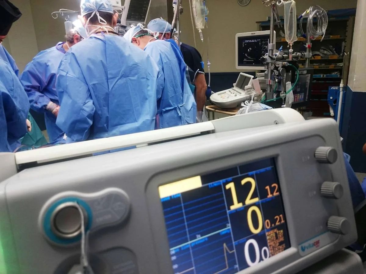 Primer plano de un monitor médico con pantallas digitales en funcionamiento dentro de un quirófano con cirujanos de fondo.