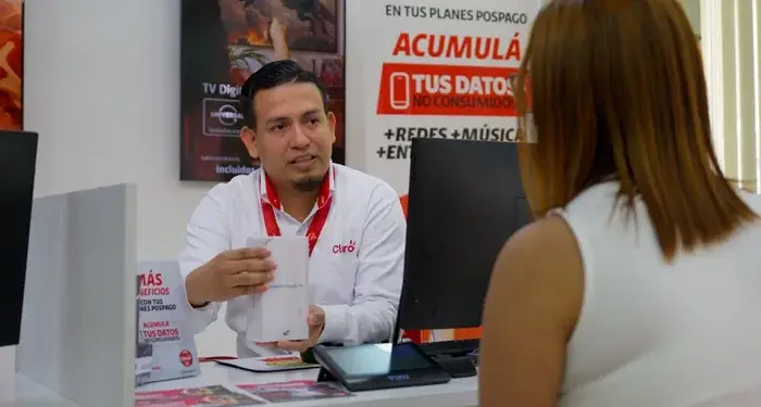 Asesor de Claro explica plan pospago a clienta en oficina. Muestra un producto, con carteles de servicios móviles y TV digital de fondo.