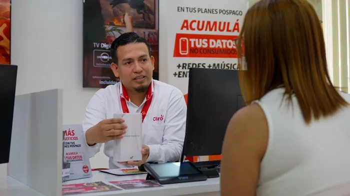 Asesor de Claro explica plan pospago a clienta en oficina. Muestra un producto, con carteles de servicios móviles y TV digital de fondo.